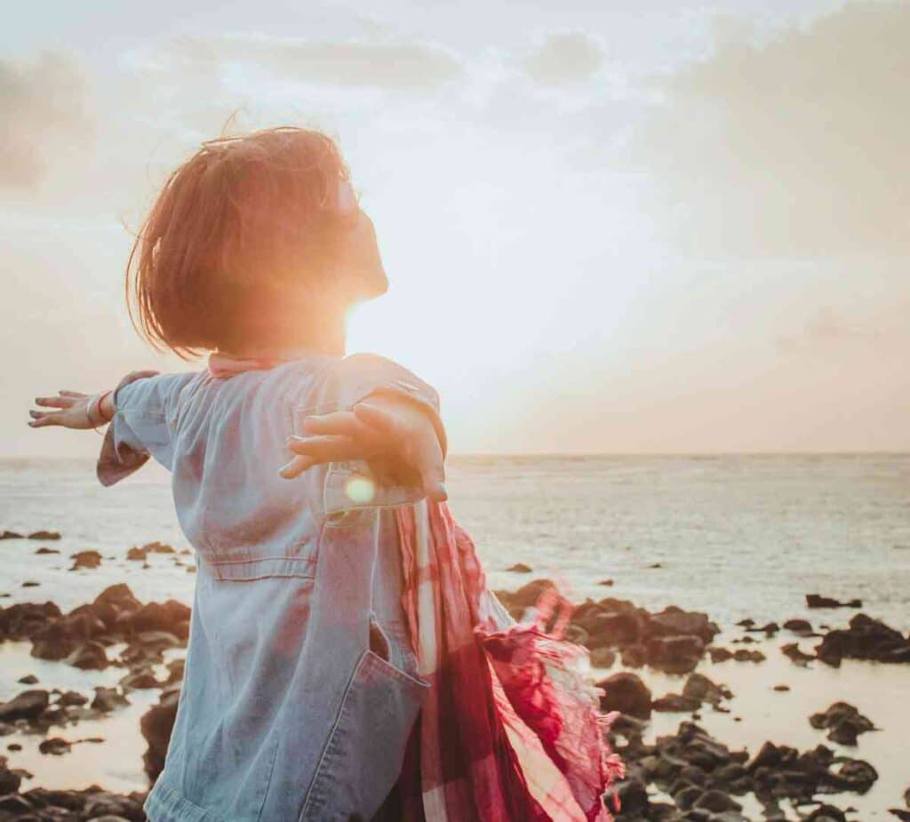 Woman standing by the ocean with arms open toward the sun, symbolizing freedom and calm after anxiety therapy.