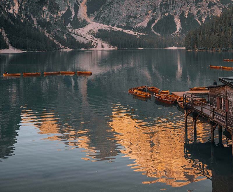 A sunlit mountain is reflected in a calm lake with wooden boats.